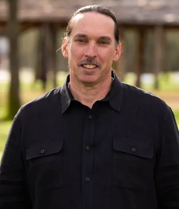 Allan Campbell standing outdoors in a black button-up shirt, smiling slightly with trees and a wooden arbor behind him.