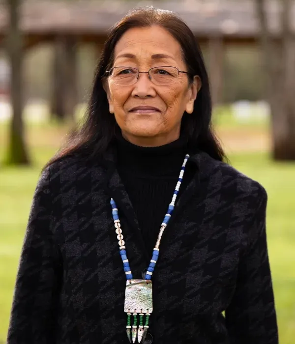 Annie Davis wearing glasses and a traditional beaded necklace, standing outdoors with a calm expression.