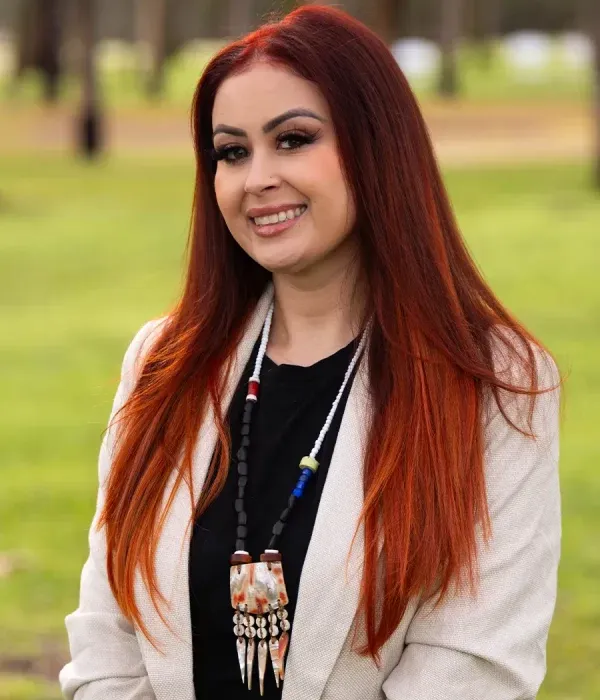Jessica Jimenez smiling outdoors in a light blazer with long red hair and a beaded necklace.