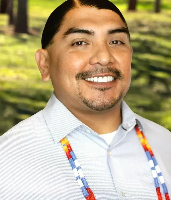 John Williams smiling in a light button-up shirt with a colorful beaded lanyard, standing against a blurred outdoor background.