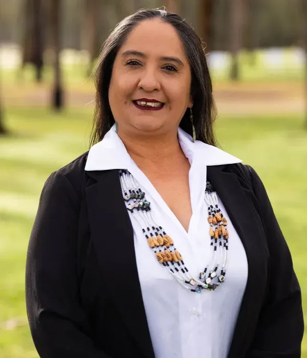 Regina Cuellar smiling outdoors, wearing traditional beaded necklaces and a black blazer over a white shirt.
