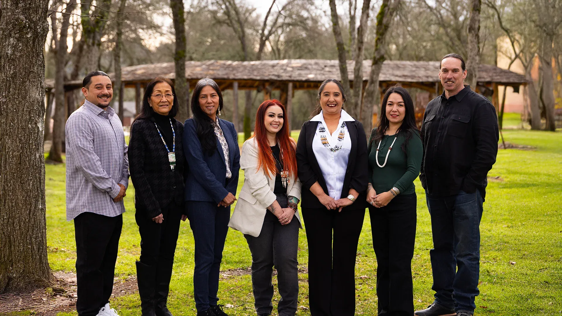 Eight members of the Shingle Springs Tribal Council standing outdoors in a grassy area with trees and a wooden arbor in the background.